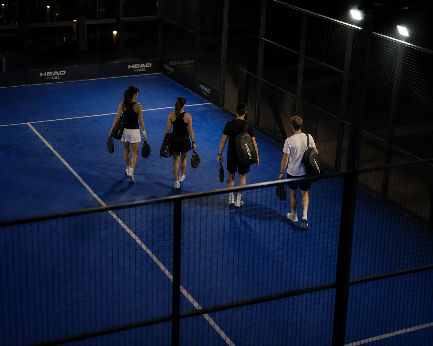 Four players walking onto a lit court at night, seen from above through the fence. Nobody has served yet. The whole site has been about getting to this moment.